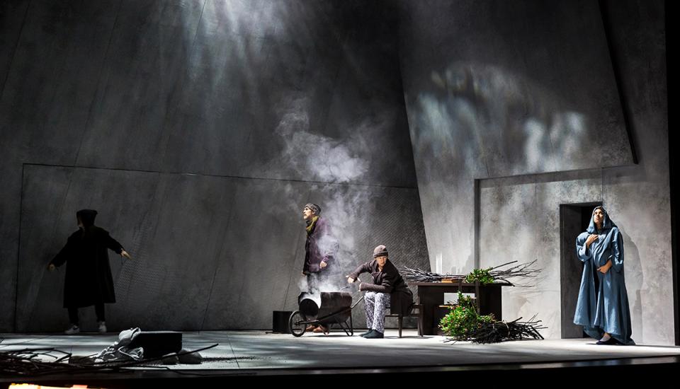 Un personnage fait du feu au centre d'une pièce aux murs gris. De la fumée s'élève. À droite, une femme en bleu tient un bébé. Près du feu, un homme reste debout, regardant à gauche. À gauche, un personnage n'est vu que de dos.